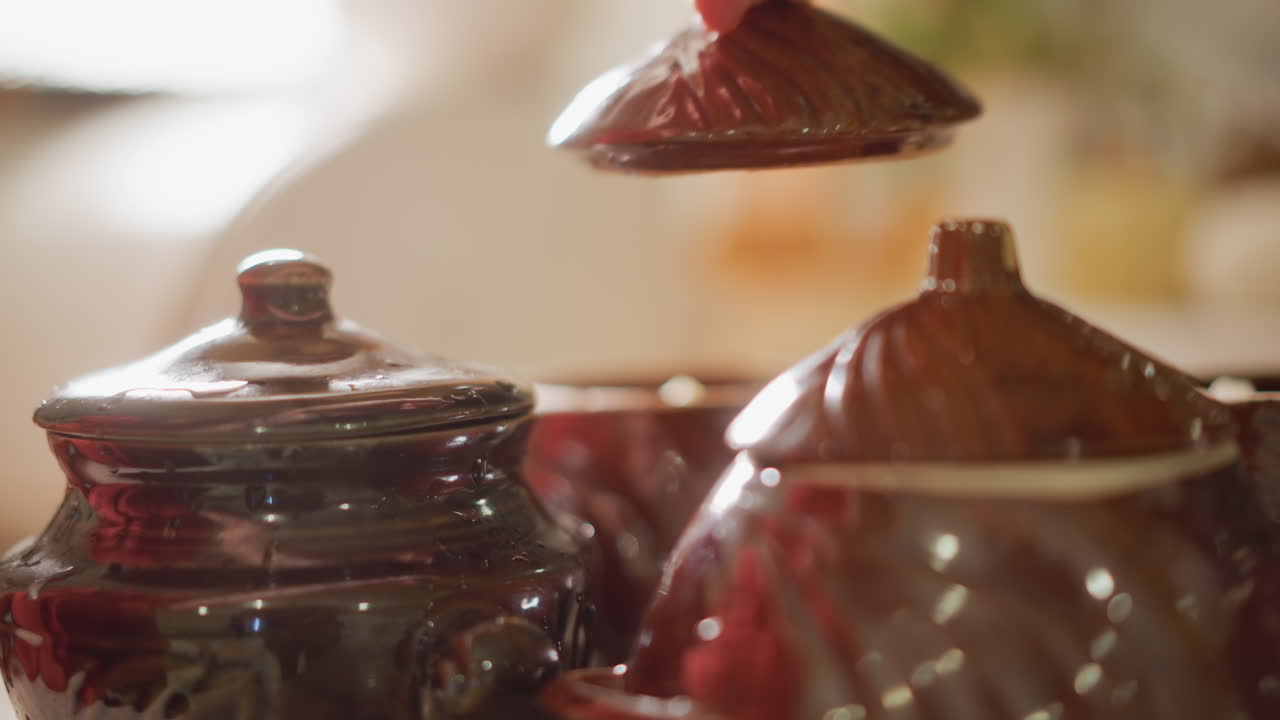 Closeup hands placing ceramic lid on glazed clay pot on wooden counter, warm sunlight highlights glossy finish and textured ridges, slow deliberate motion, intimate home cook preparing stew