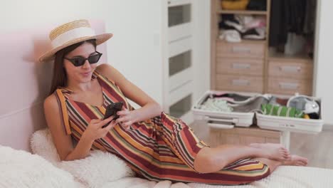 hermosa chica con gafas y sombrero elige viajar por teléfono a dónde ir de vacaciones, una maleta en el fondo recogida