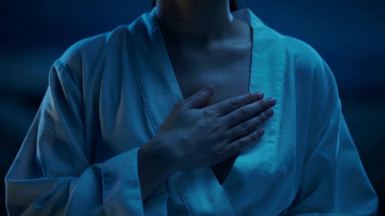 Posizionando la mano destra, donna che si sistema sul petto sinistro e sulla clavicola in una stanza illuminata di blu, indossando una vestaglia