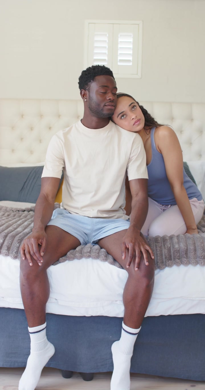 Couple sitting on bed with tufted headboard, woman shifting closer and leaning, seeking comfort