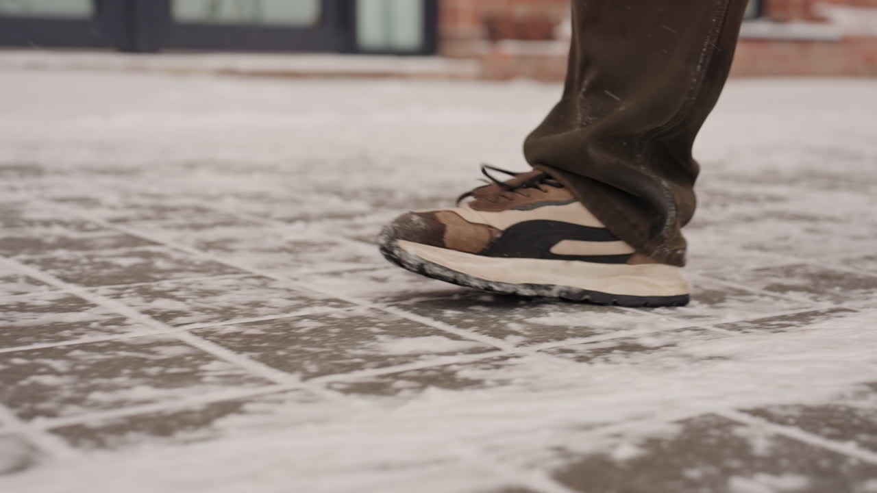 Closeup snowy sneaker steps on icy sidewalk lowangle shots capture cautious stride, textured tiles dusted with fresh snow, brown sneakers with rubber soles, cuffed trousers, quiet early morning urban
