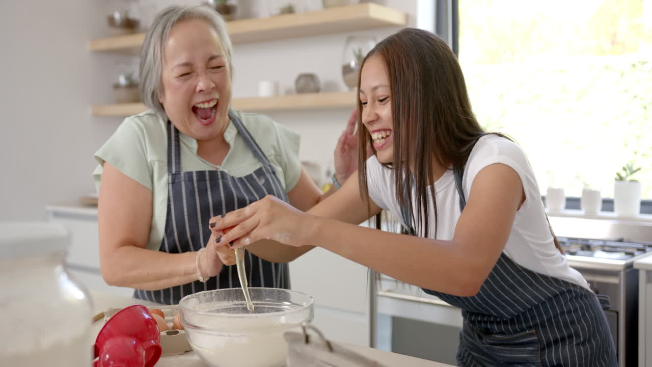 Laughing together, asian grandmother and granddaughter baking while cracking eggs in kitchen