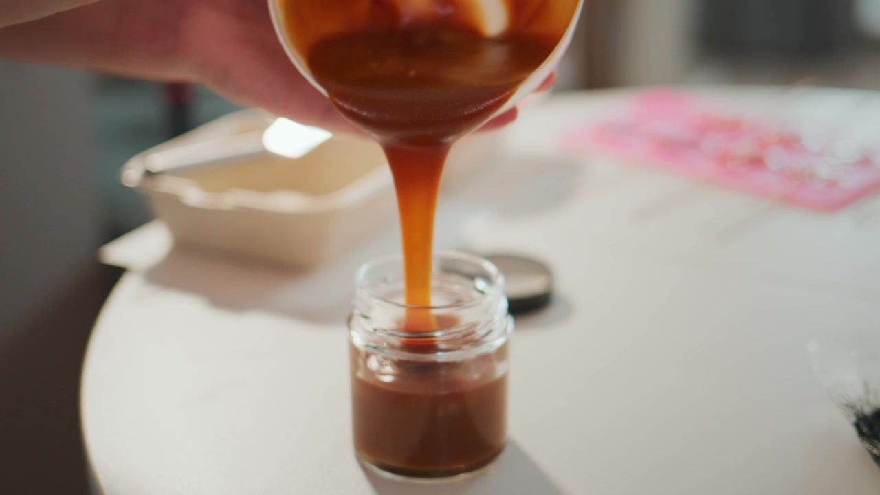 Una mano versa sciroppo di caramello denso in un barattolo di vetro. Il liquido liscio e dorato scorre lentamente. Lo sfondo mostra un bancone da cucina con carta rosa. Perfetto per blog di cucina, video di ricette o pubblicità di dolci.