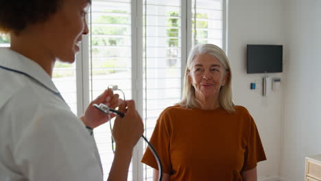 female nurse wearing uniform listening to senior female patient's chest in private hospital room