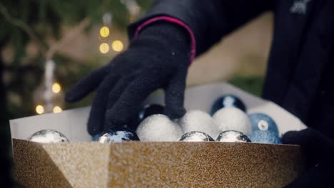 blue, white and silver reflecting spheres in a box grabbed by a hand in a woolen glove during christmas