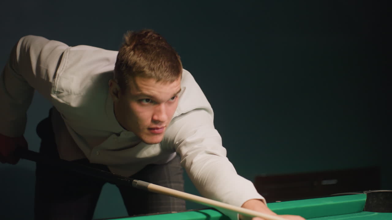 Close-up of sports enthusiast in white shirt leaning over green pool table, gripping cue stick with intense focus. Eyes locked on cue ball as he prepares to take shot, demonstrating precision, skill
