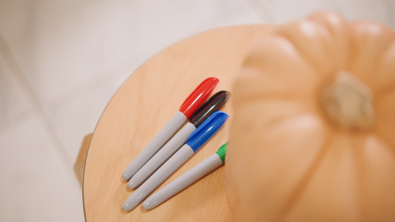 Overhead pumpkin on wooden stool markers arranged nearby, topdown view showing red blue and green marker caps, soft warm light, shallow depth of field, neutral beige background, calm autumn mood ideal