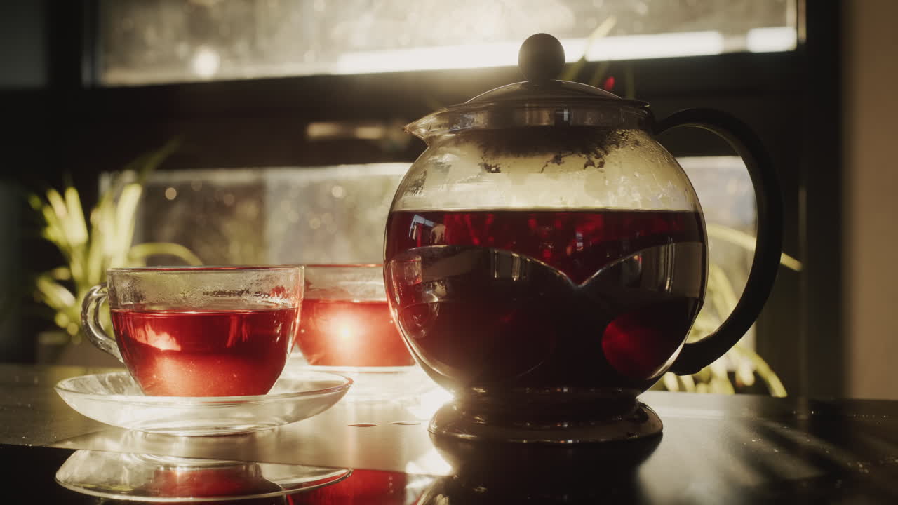 A glass teapot with tea and two cups stand by the window from which the sun shines.