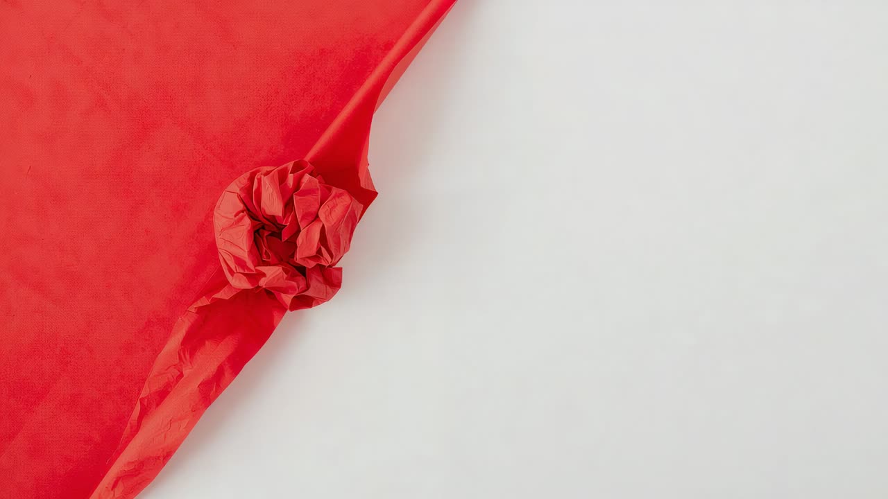 Déroulement hors cadre d’une main tirant du papier de soie rouge sur une table blanche, lissant un nœud froissé