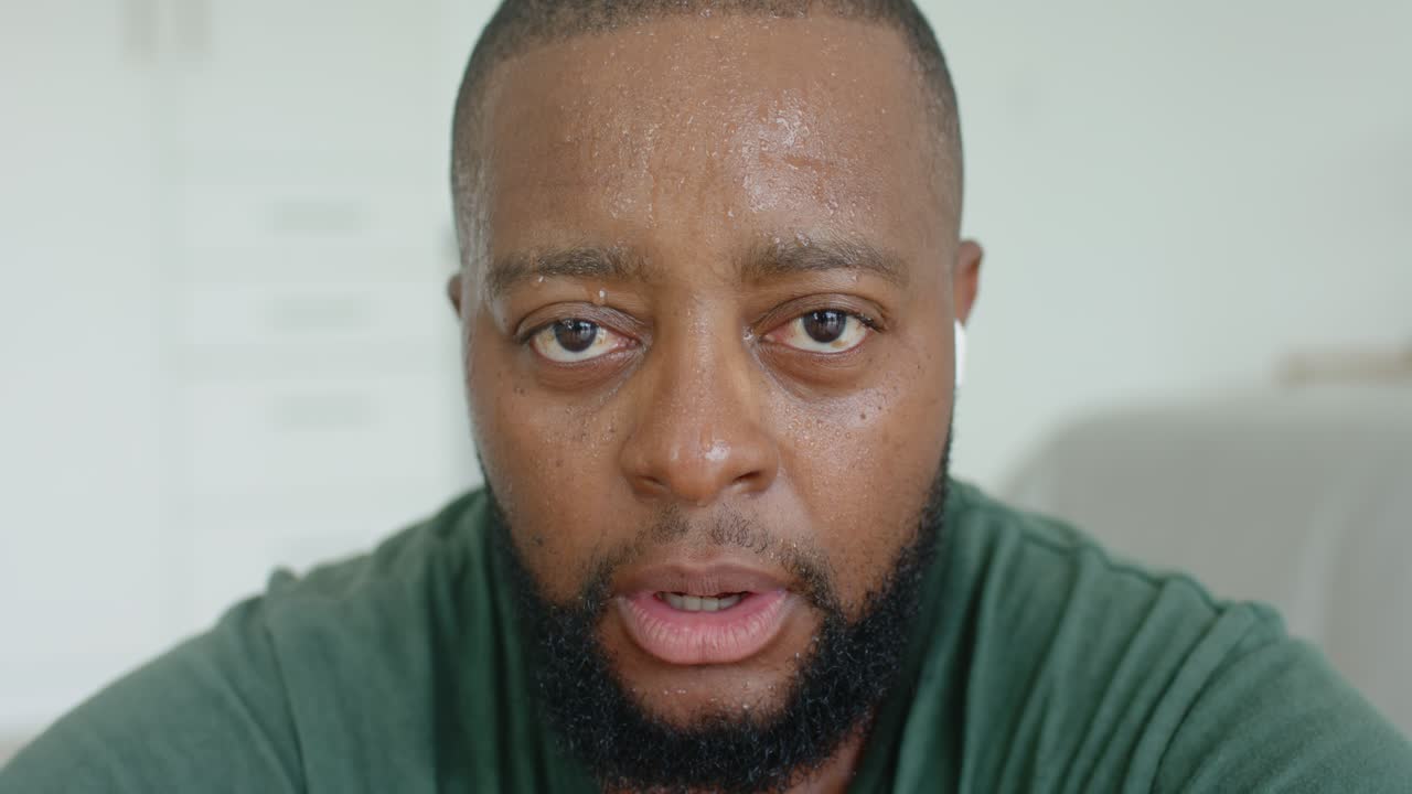 Uomo adulto africano rivolto verso la telecamera, che respira pesantemente dopo uno sforzo, in salotto con un auricolare bianco.
