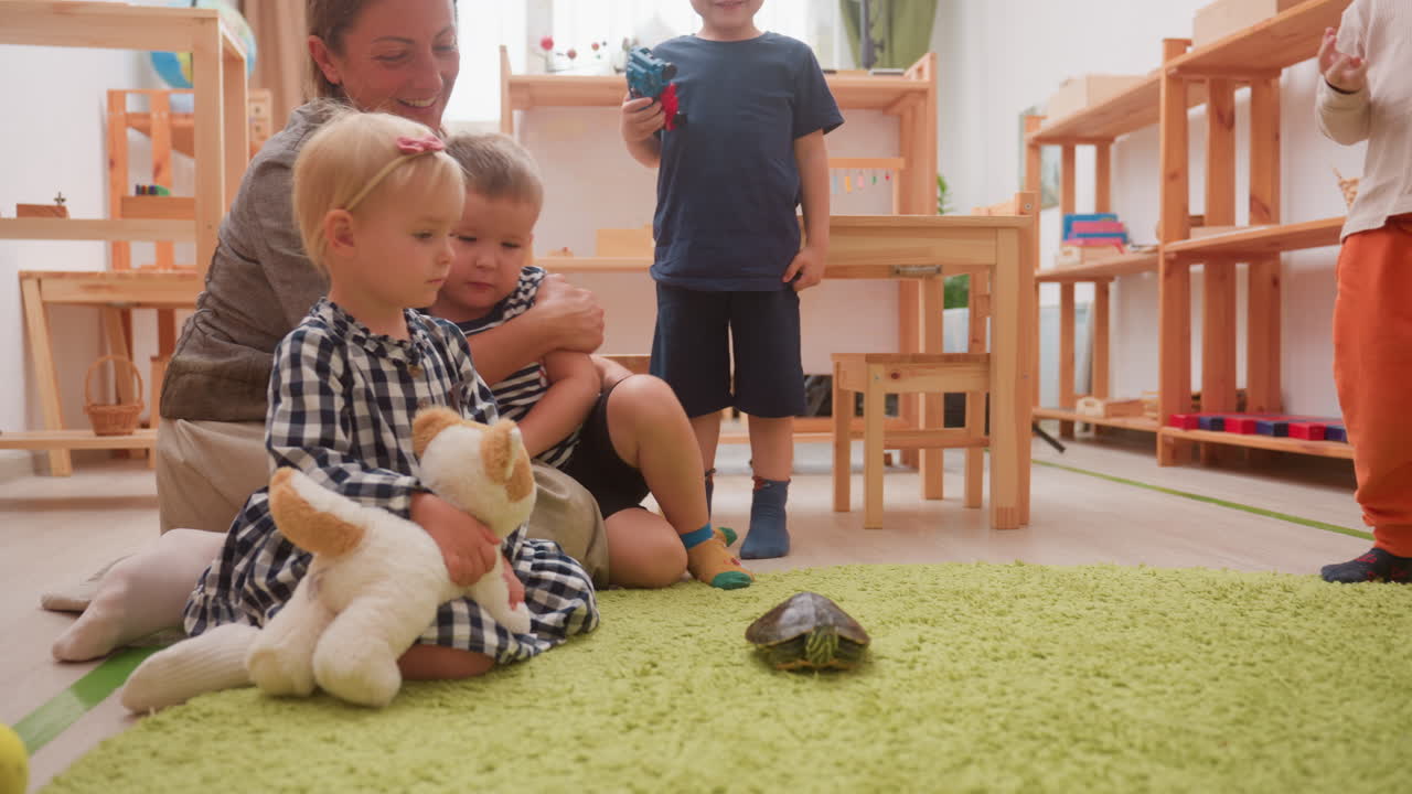 BetreuerIn mit Minderjährigen im Klassenzimmer, die eine Schildkröte auf einem grünen Teppich beobachten, während sich ein Junge mit Hörgerät der/dem BetreuerIn nähert, während ein Mädchen, das ein Stofftier hält, und ein anderer Junge mit einem Spielzeugauto aufmerksam zusehen