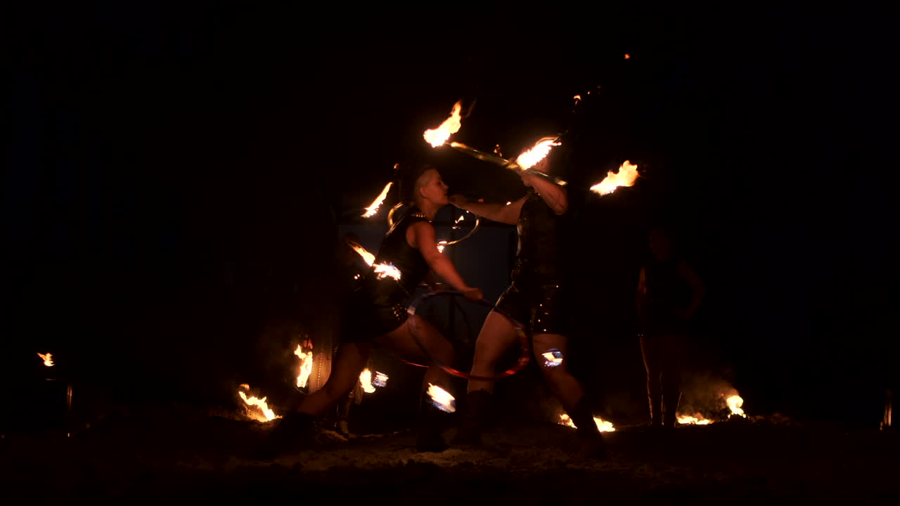 Slow motion: Professional fire performance three women in dripping clothes dance and spin with flaming torches and a man with flamethrowers in the background.