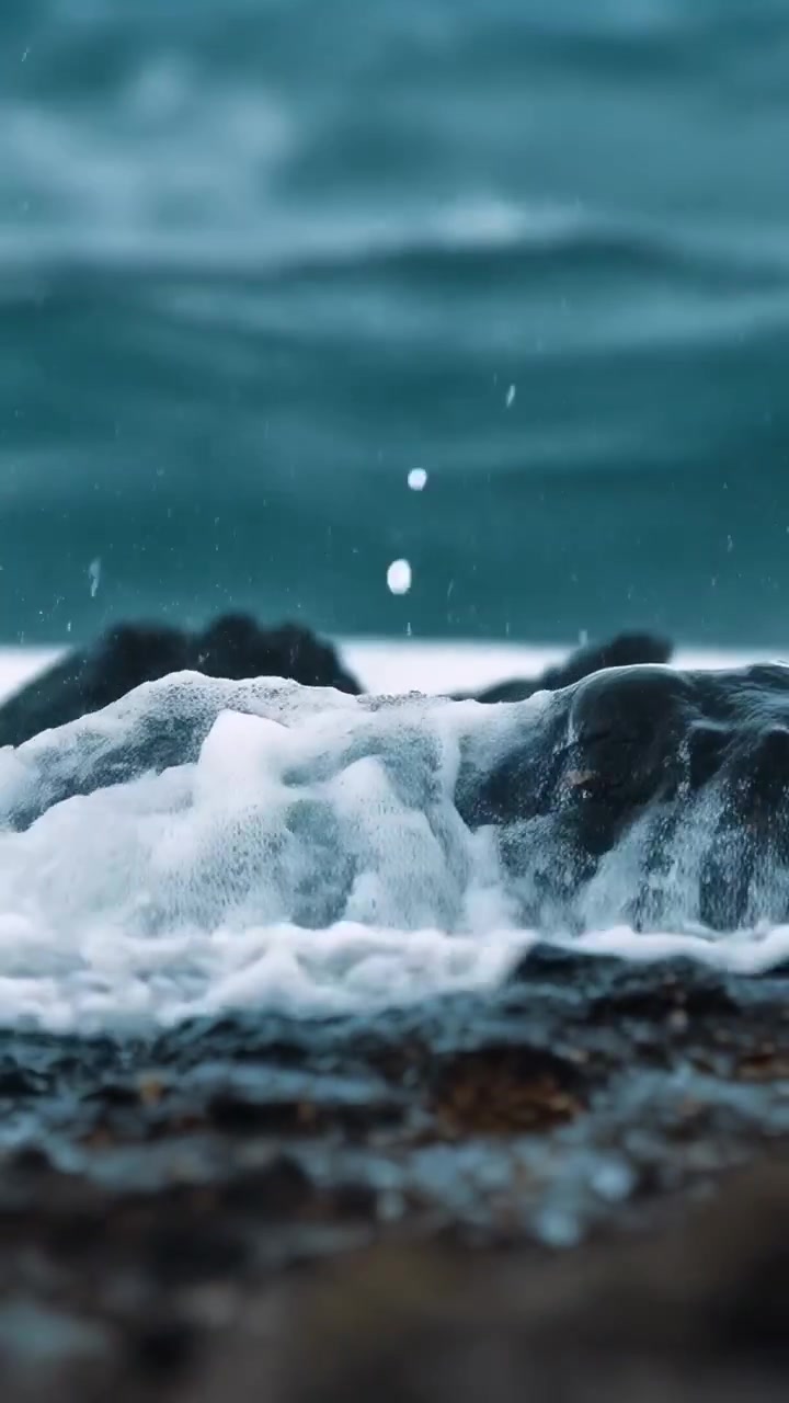 Video verticale: Onda che si infrange violentemente contro una roccia scura sulla battigia, la mareggiata che solleva spruzzi e schiuma bianca