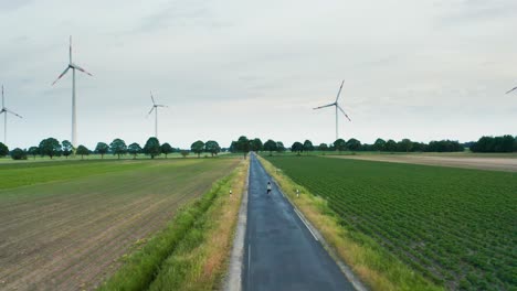 Una-Joven-Está-Montando-Su-Bicicleta-En-Un-Largo-Camino-A-Través-De-Un-Campo-Verde-De-Molinos-De-Viento-Parque-De-Tecnología-De-Energía-Renovable-Con-Turbinas-Altas-En-Tierras-De-Cultivo-Holandesas-4k-Después-De-Un-Disparo-De-Drones