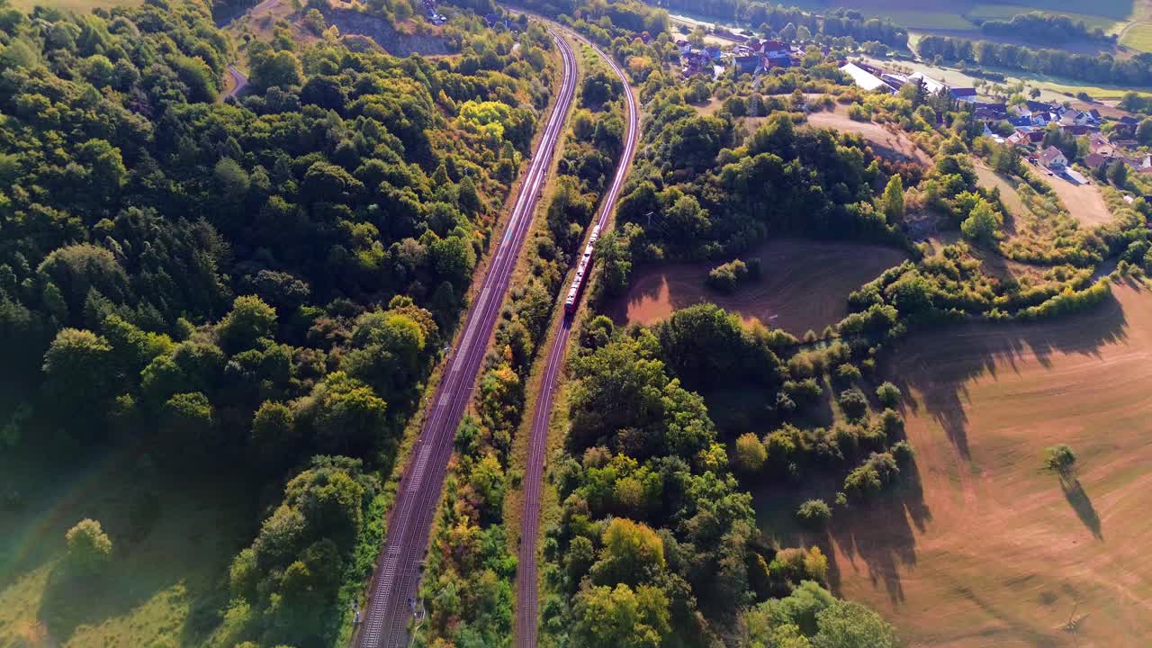 Aerial drone view of passenger train moving along railway tracks through forest and farmland with rural village in distance