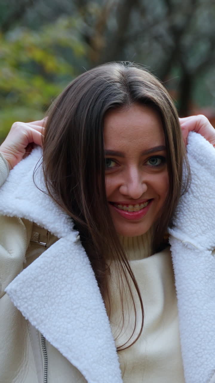 Cheerful positive lady puts on warm jacket walking in the park. Woman looks into camera smiling posing at the backdrop of beautiful autumn weather. Vertical video
