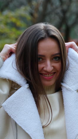 cheerful positive lady puts on warm jacket walking in the park. woman looks into camera smiling posing at the backdrop of beautiful autumn weather. vertical video