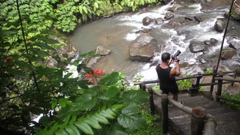 una toma rápida de izquierda a derecha de hojas mojadas y húmedas a un joven blanco vestido de negro, de pie tomando fotos de un río y una cascada debajo de unas escaleras empinadas en la jungla de bali