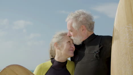 close up of a senior couple in wetsuit with surfboard standing on the sandy beach, looking at the camera and kissing