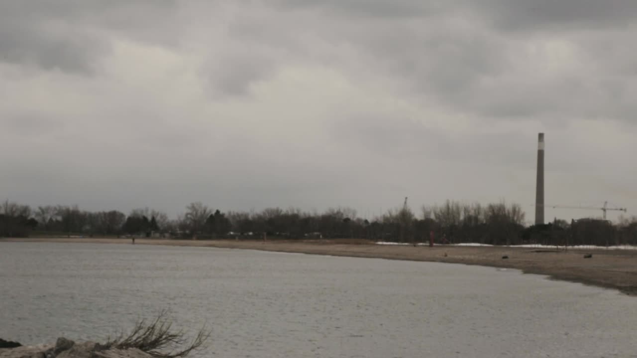 Beach In Toronto, Canada - Calm Sea Waves Splashing On The Stony Shore With Light House Towers And Dry Trees On A Cloudy Day - Panoramic Shot