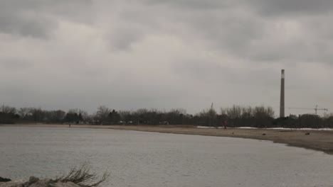 beach in toronto, canada - calm sea waves splashing on the stony shore with light house towers and dry trees on a cloudy day - panoramic shot