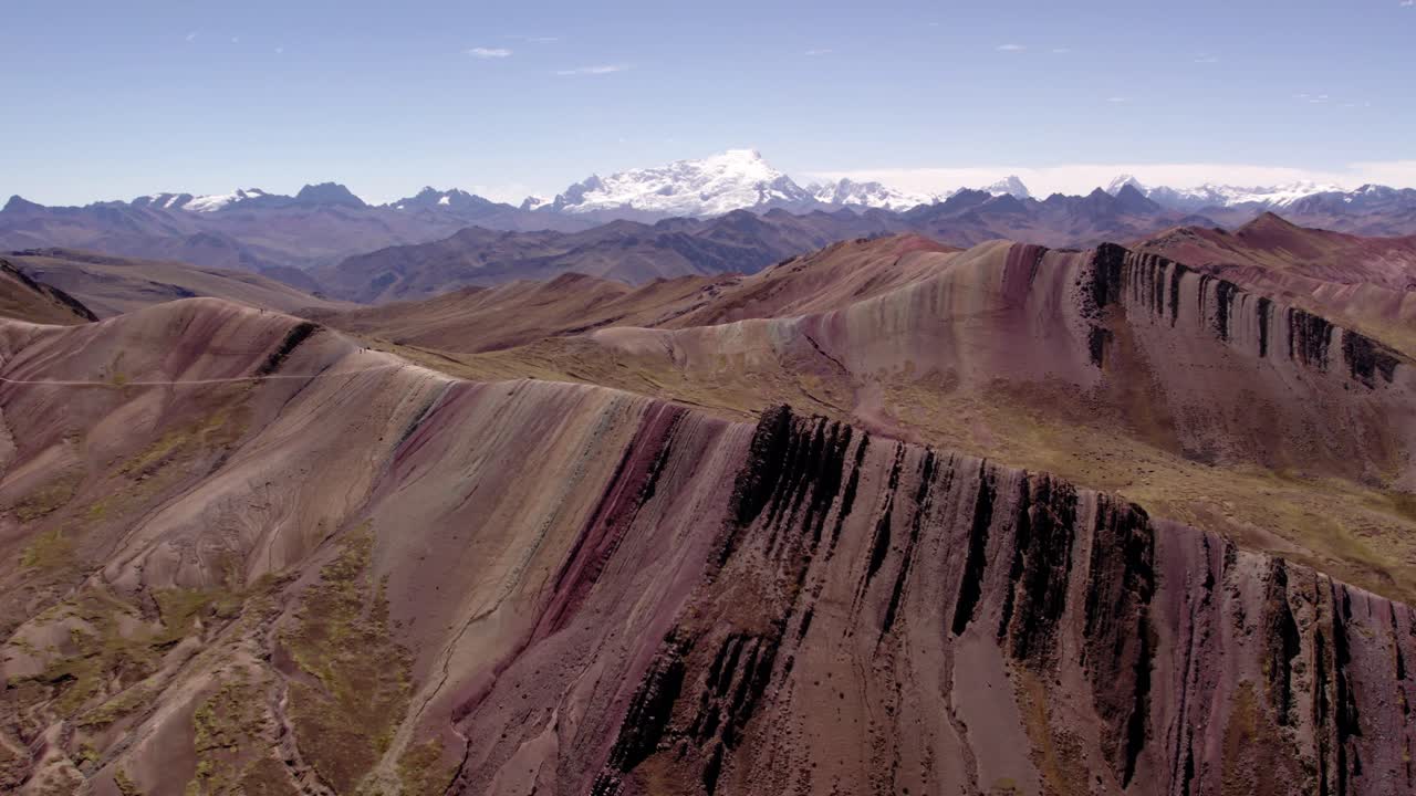 Vibrant rocky terrain stretches across the Peruvian Andes, featuring layers of earth tones. Snow-capped peaks rise in the distance with the clear blue sky above.
