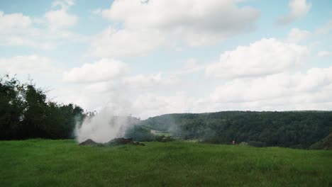 pile of earth on large green field with white smoke moved by wind