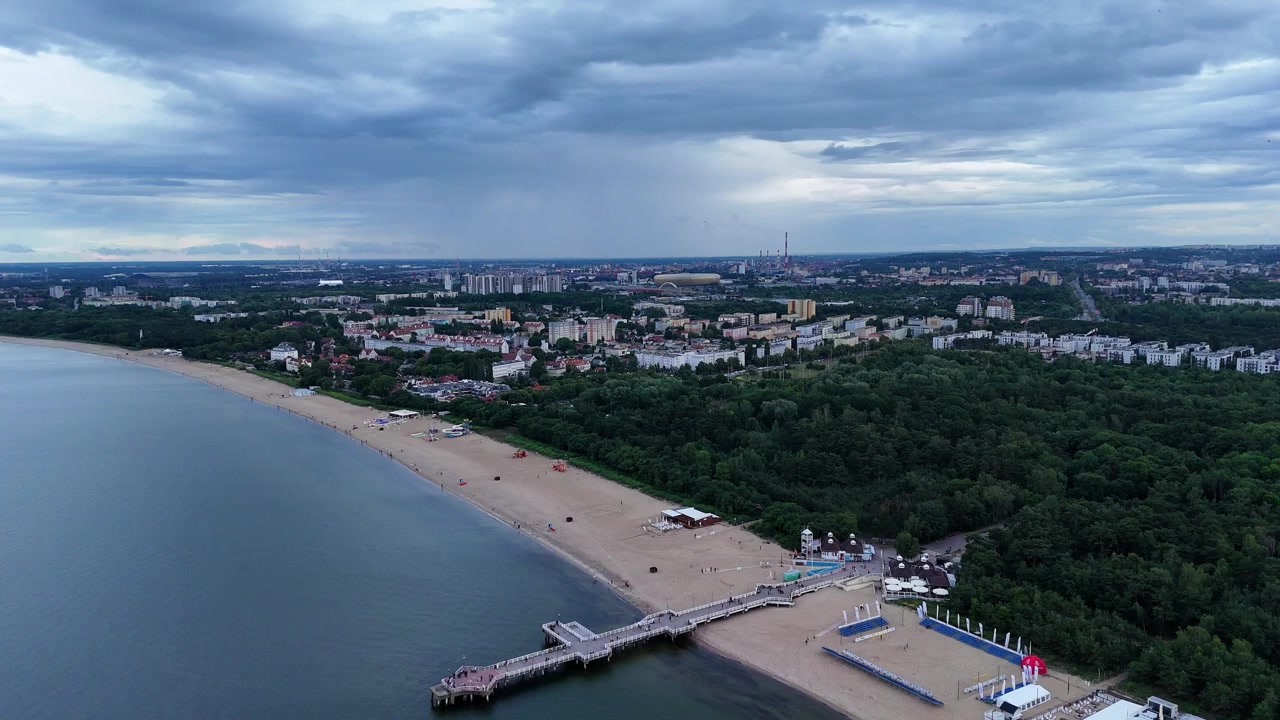 La spiaggia di Danzica si estende lungo la costa con un imponente molo che si protende nel mare. Lo skyline della città sullo sfondo presenta un mix di aree urbane e verdi sotto un cielo nuvoloso, ripresa rivelatrice con drone.