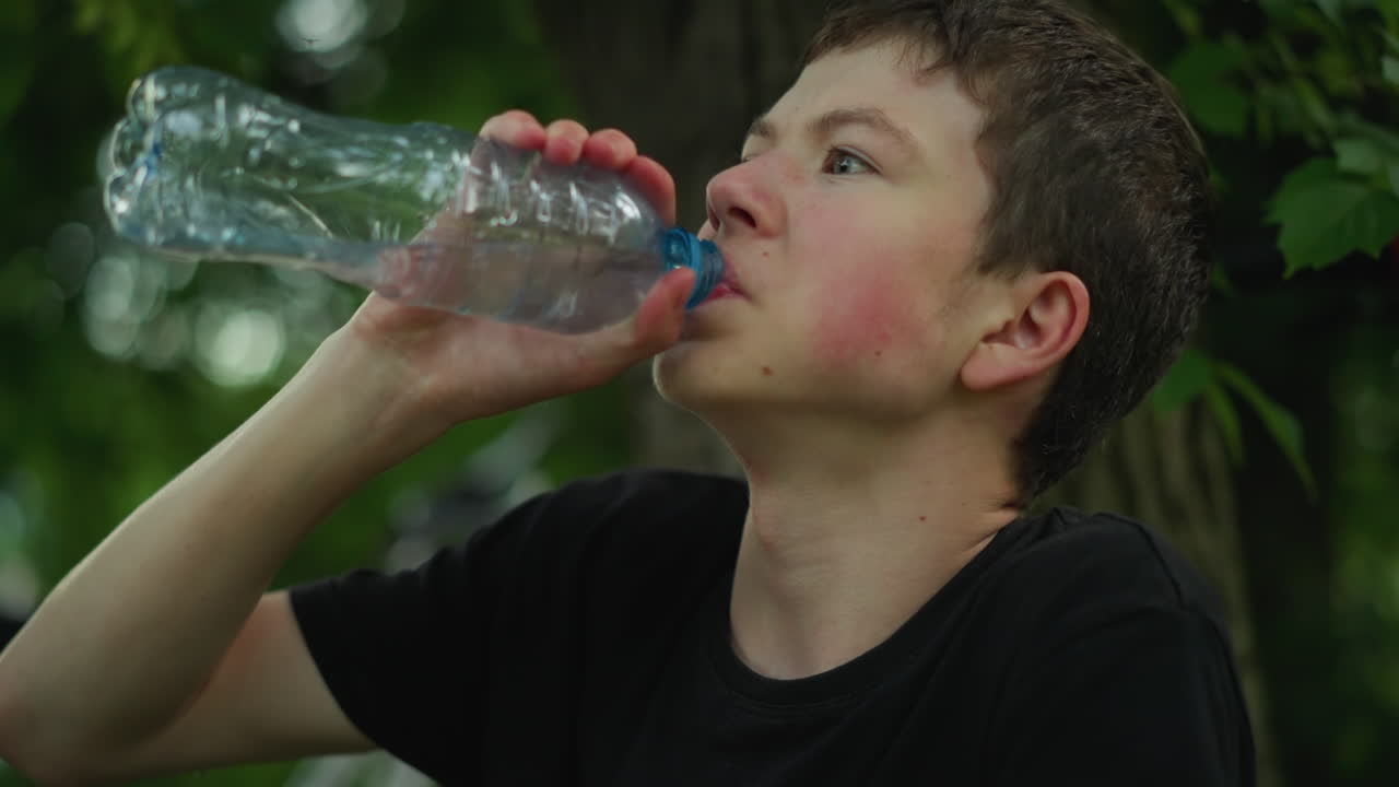 검은 티셔츠를 입은 어린 소년은 나무 에 앉아 플라스틱 병에서 물을 마시며, 따뜻한 날에 녹색으로 둘러싸인 신체 활동 후에 냉각하는 것처럼 보입니다.