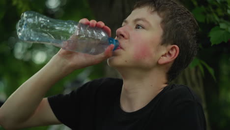 niño joven con camiseta negra bebe agua de una botella de plástico mientras está sentado bajo un árbol, parece estar enfriándose después de la actividad física, rodeado de vegetación en un día cálido