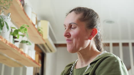 elderly woman in green clothing enjoys eating berry in cozy kitchen with wooden shelves and potted plants, smiling with satisfaction as she savors healthy fruit in peaceful home environment