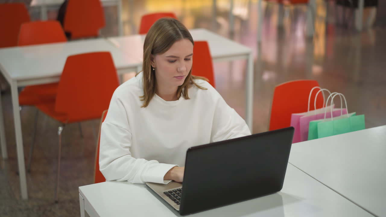 mujer joven sentada en un restaurante moderno trabajando en una computadora portátil, mirando lejos pensativo con bolsas de compras en la mesa, interior vibrante en el fondo