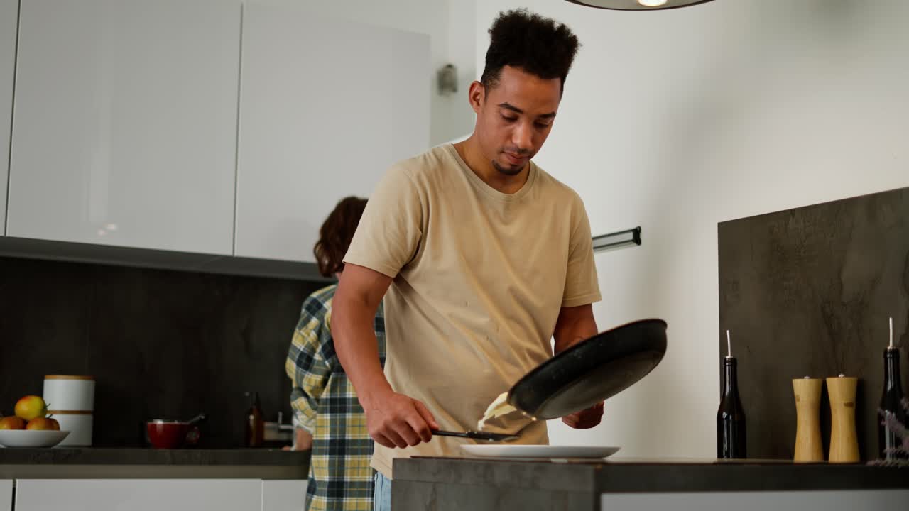 un joven moreno de piel negra con una camiseta beige pone huevos revueltos en un plato mientras están preparando el desayuno junto con su novia un joven adulto moreno que lava los platos que están preparando para el desayono y por la mañana en un apartamento moderno