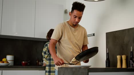 a young black-skinned brunette man in a beige t-shirt puts scrambled eggs on a plate while they are preparing breakfast together with his girlfriend a young adult brunette who washes the dishes they are preparing for breakfast and in the morning in a modern apartment