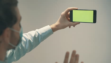 hombre con mascarilla sosteniendo un teléfono inteligente de pantalla verde con una videollamada de negocios