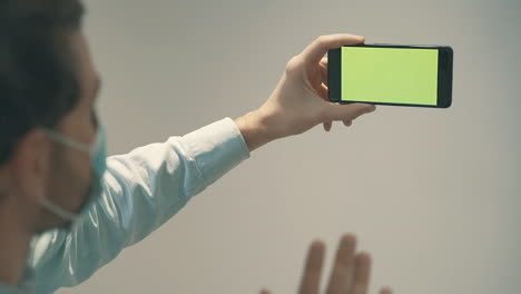 male with face mask holding a green screen smartphone having a business video call