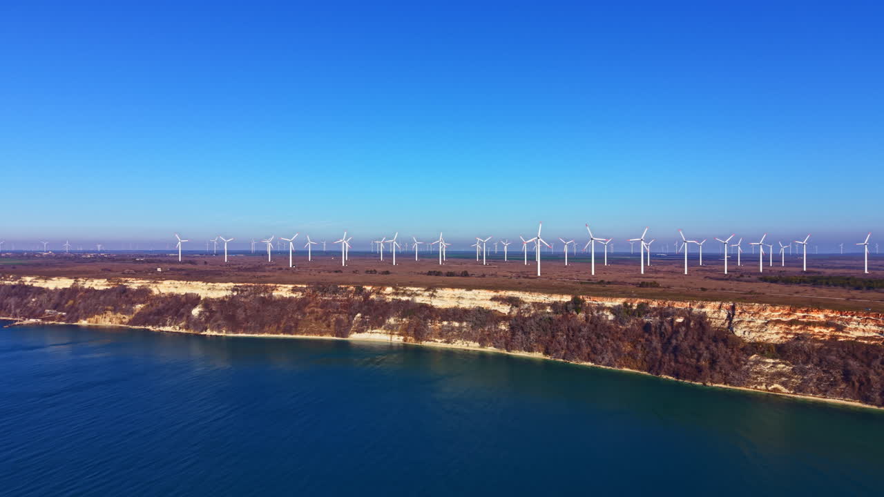 Wind turbines stand along the coast under clear blue sky near the water while the land rises into cliffs in a landscape of nature with renewable energy