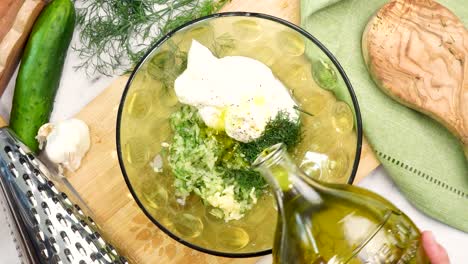 making tzatziki greek or mediterranean sauce or dip for a party. dip or sauce in a green bowl on a wooden cutting board with dill, green napkin and a grater. adding olive oil to the tzatziki sauce.