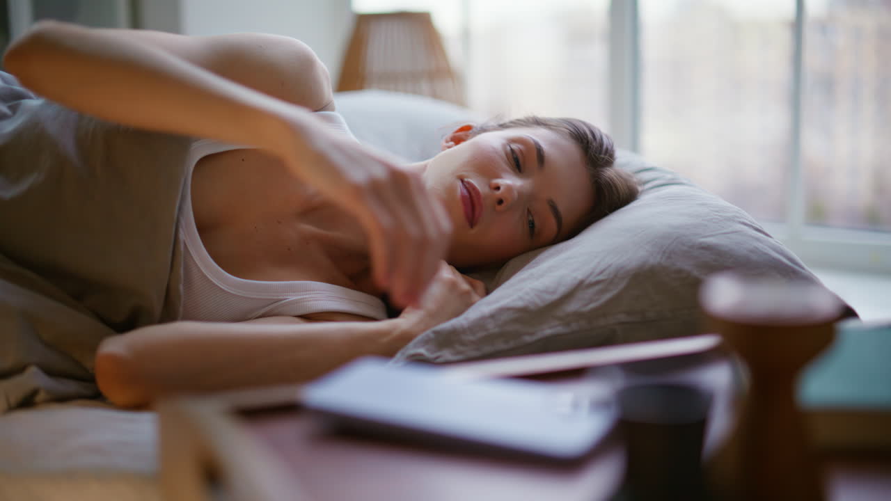 Morgenkvinde ligger i sengen, vågner op, nærbillede. Glad pige kigger på mobiltelefon sms