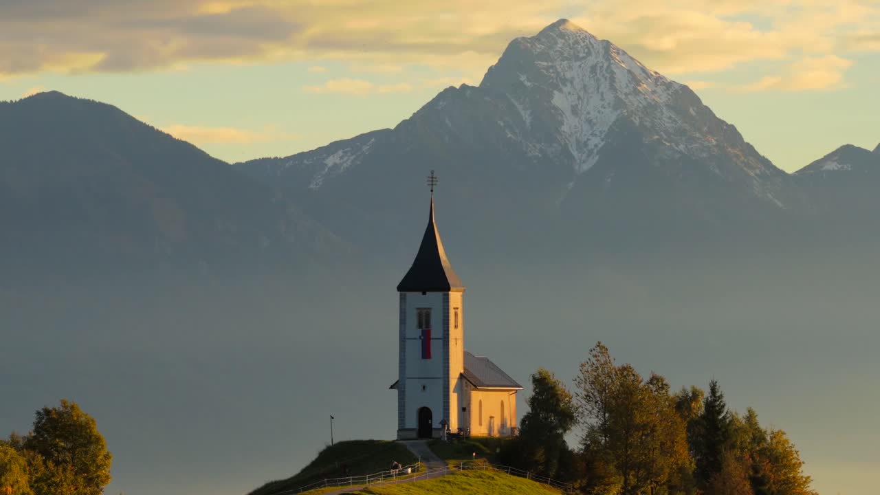 Aerial push-in shot of the Church of St. Primus and Felician at Jamnik, Slovenia, surrounded by autumn hills and backed by the Julian Alps, showcasing peaceful alpine scenery at sunrise