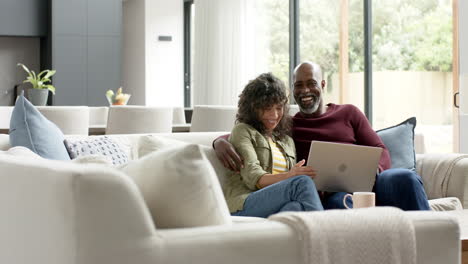 Happy-biracial-couple-sitting-on-couch-and-using-laptop-at-home,-slow-motion