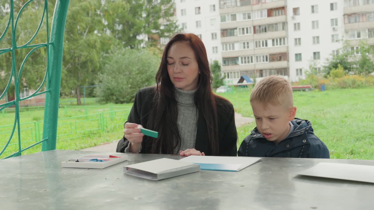 Tutoraggio al tavolo all'aperto per la preparazione scolastica: una madre caucasica funge da tutor, mentore e coach di studio, guidando un bambino attraverso esercizi sul quaderno, indicando le pagine e incoraggiandolo a prendere appunti e a fare pratica.