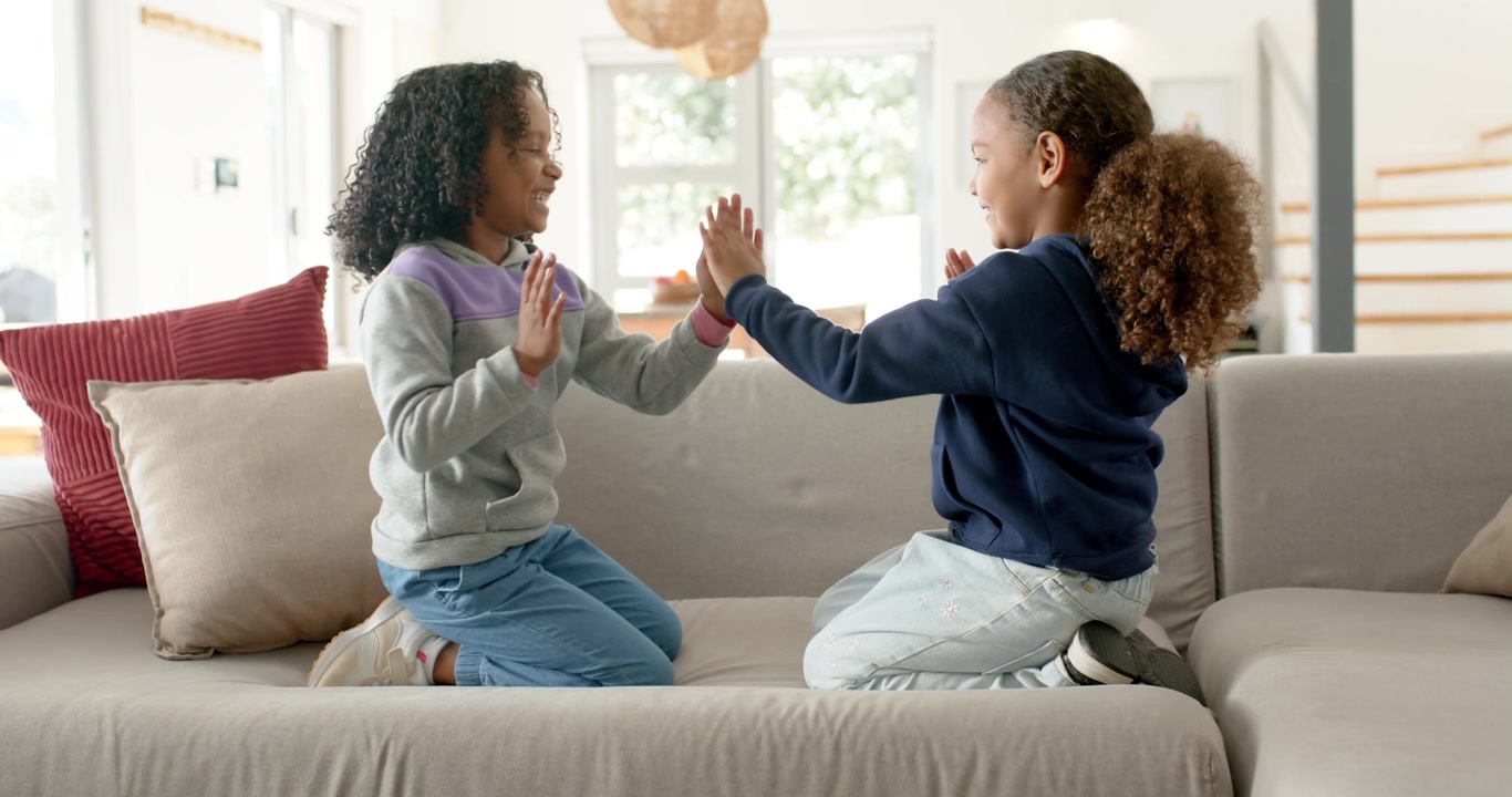 African American youth girls kneeling on couch at home raising palms clapping, high-fiving