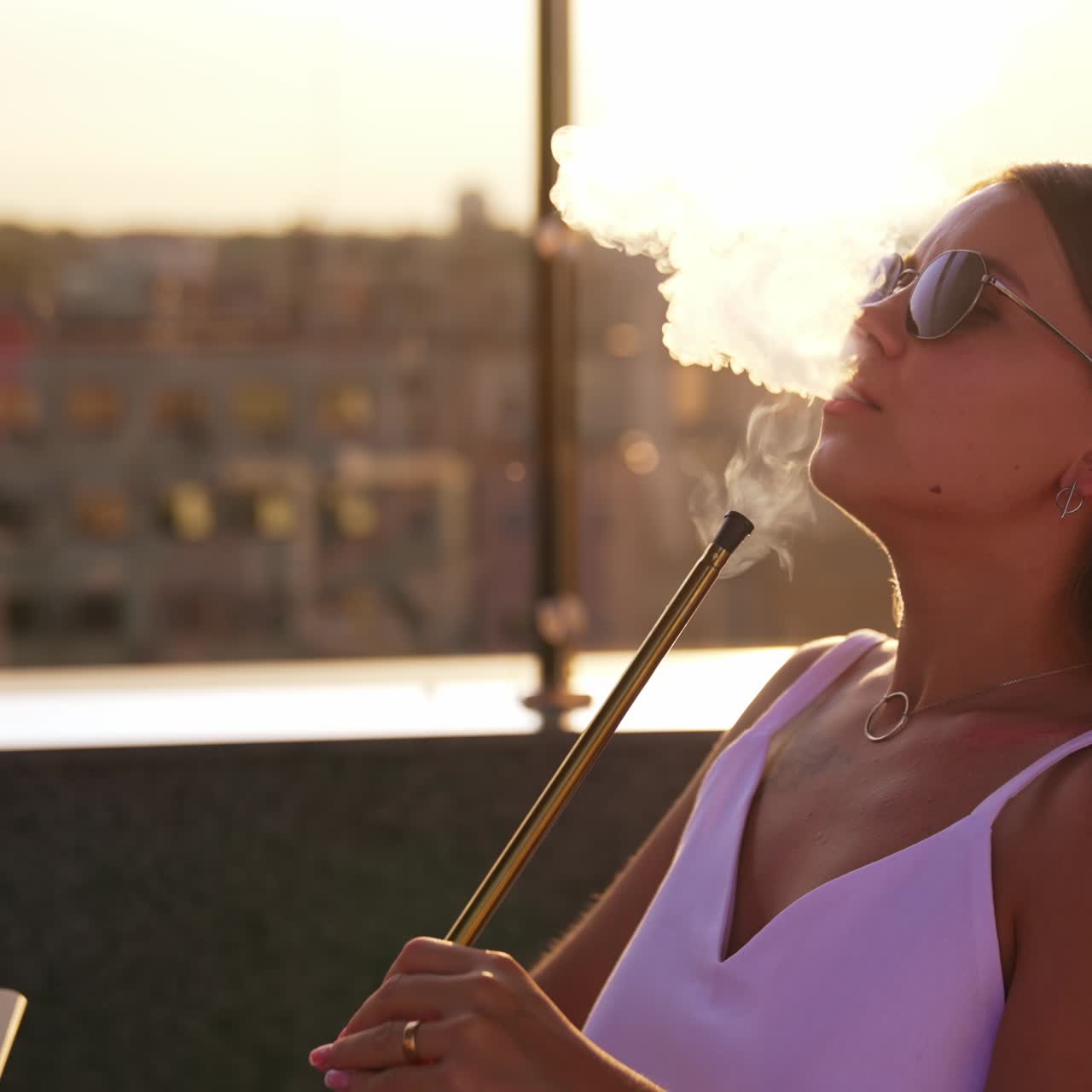 Blonde girl in sunglasses takes a pipe of a hookah to her lips. Woman makes smoke into air in sun rays. Blurred backdrop