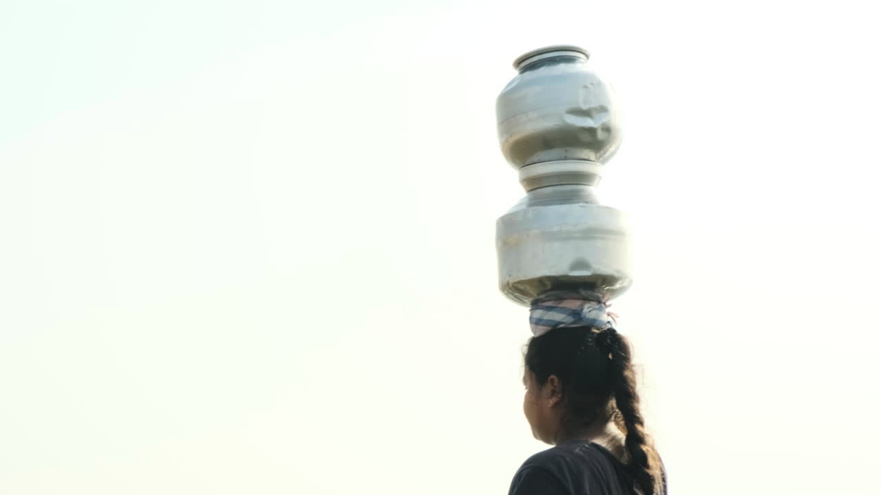 A slow-motion long mid shot of a middle aged Indian lady carrying two metal pots on her head and walking in broad sunny daylight