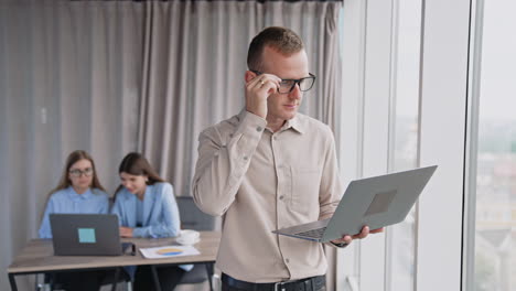 mid aged man wearing glasses holding laptop working on it standing at the window. female colleagues working at computer at backdrop in blur.