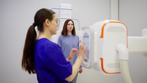 A-confident-brunette-girl-doctor-with-glasses-in-a-blue-uniform-makes-fluorography-of-a-young-brunette-girl-in-a-blue-jacket-in-the-office-of-a-modern-clinic