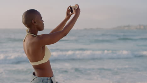 portrait of attractive african american woman using smartphone taking photos on mobile phone camera enjoying beautiful seaside beach wearing sexy bikini slow motion