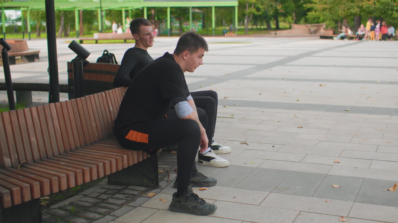 Dwóch mężczyzn rozmawia na drewnianej ławce, w słoneczny dzień na miejskim placu z drzewami; jeden pochyla się do przodu, a drugi słucha i oferuje ciche wsparcie, ciepły empatyczny nastrój i delikatna szczera wymiana zdań, otwarta