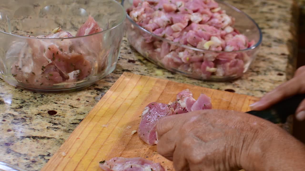 Close-up tangan wanita lansia sedang memotong daging mentah yang sudah dibumbui menggunakan pisau tajam. Latar belakang dengan lebih banyak potongan di mangkuk kaca terdekat